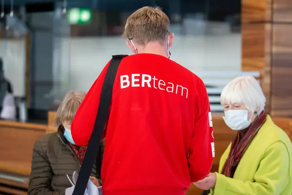 A BER airport employee wearing a red "BERteam" sweatshirt talks to two women with face mask