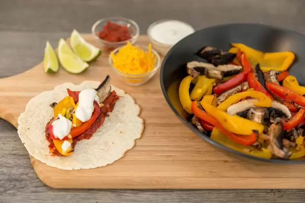 A fajita with hot tomato sauce, peppers, mushrooms, cheddar cheese and cream with lime on a wooden cutting board next to a black bowl