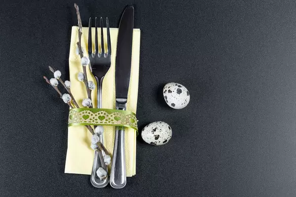 A fork and knife with a yellow napkin , willow branches and quail eggs on a black background. The concept of preparation for Easter (Flip 2020)