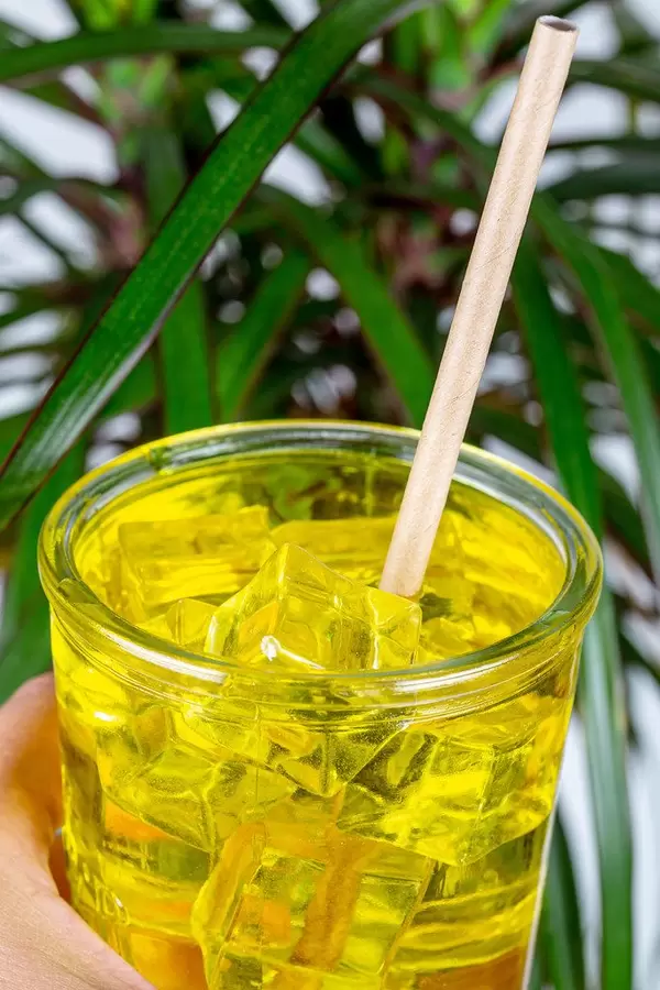 A glass of cold drink with passion fruit juice and ice cubes in a female hand on a background of green palm leaves