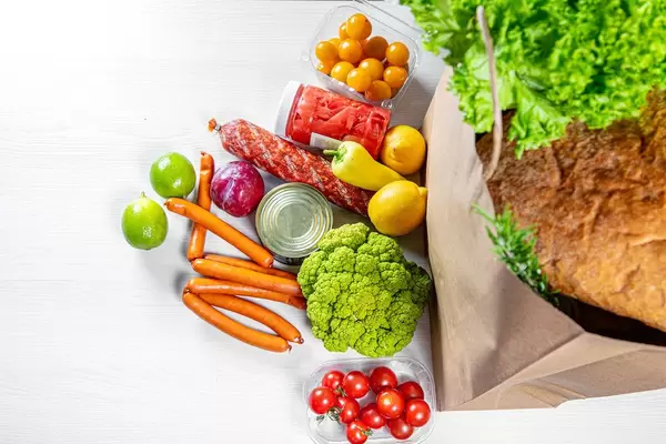 A paper bag full of groceries, surrounded by of fruits, vegetables, sausages and canned goods