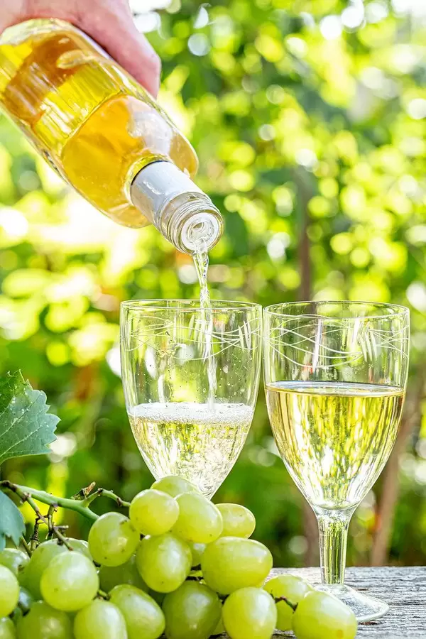 A Person is pouring White Wine into Wine Glasses with Fresh Grapes on a Wooden Table in the Foreground