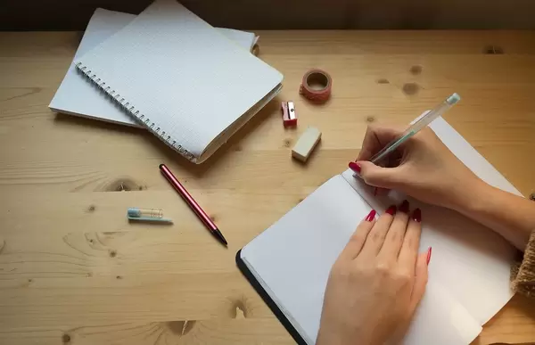 A Person writing in a Notebook with Pencil, Ereaser and Sharpener on a Wooden Table