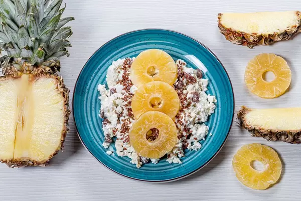 A plate of cottage cheese with flax seeds and dried fruits on a white wooden background with pieces of fresh pineapple (Flip 2019)