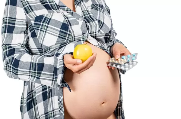 A pregnant woman holds an apple in one hand and medicines in the other