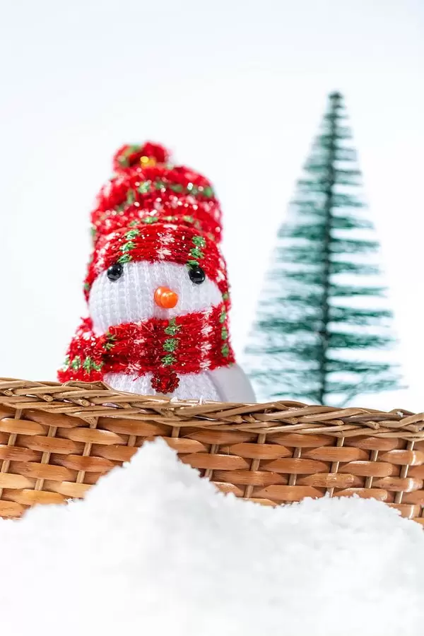 A snowman with hat and scarf inside a basket in the snow with christmas tree in the background