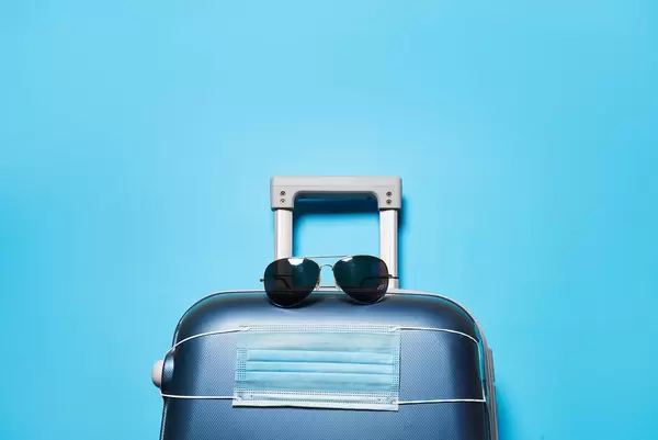 A suitcase with sunglasses and protective medical mask over blue background with copy space