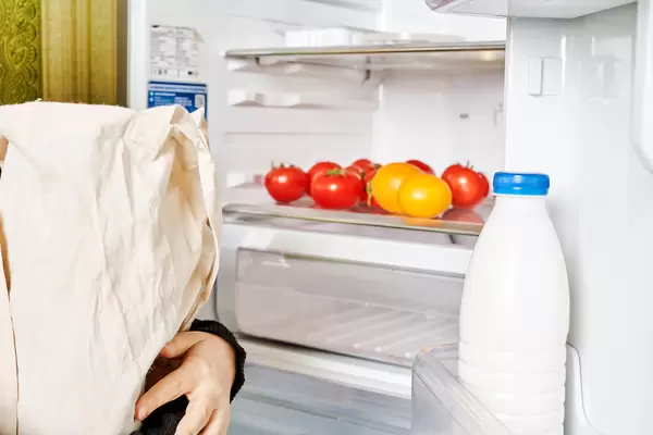 A woman holds a shopping bag full of foodstuff near the open fridge
