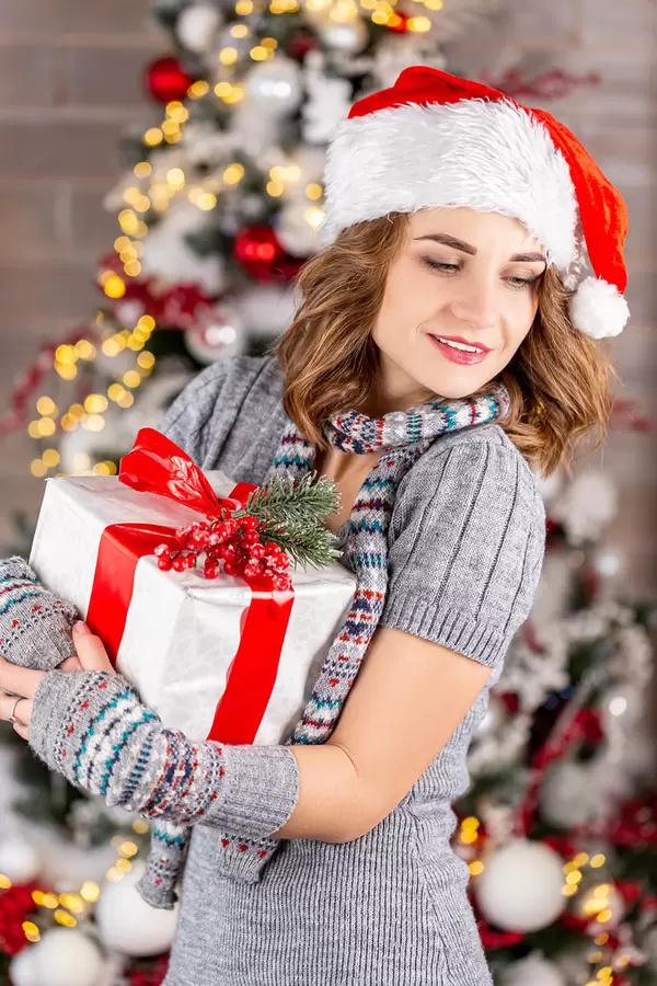 A woman with a gift in her hands in a dress, scarf, and santa hat on the background of a christmas tree