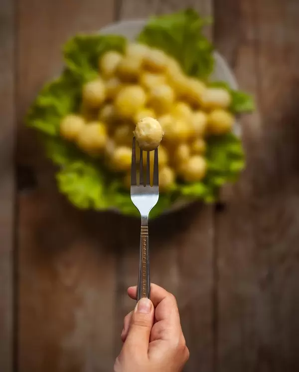A woman's hand holding cleaned fresh new potato with a fork. Full plate of fresh potatoes at the background