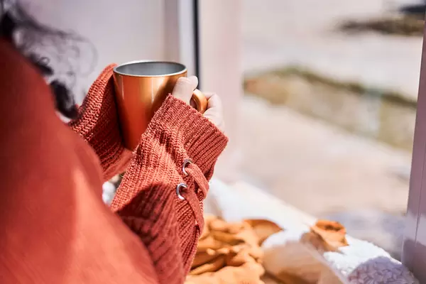 A young woman enjoying hot coffee and looking outside to autumn day near the windows