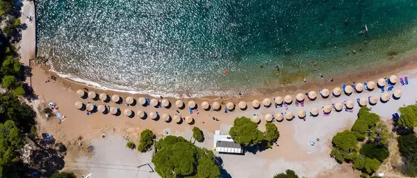 Aerial photography of tourists relaxing on vacation & swimming in the green sea at Plaza Paraskevi beach in Agii Anargiri, Spetses