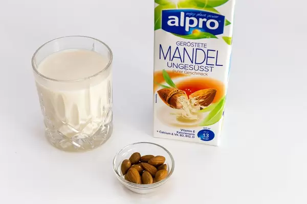 Alpro Mangelmilch, ungesüßt, in der Verpackung, neben einem Glas vegane Milch und einer kleinen Schüssel mit ganzen Mandeln