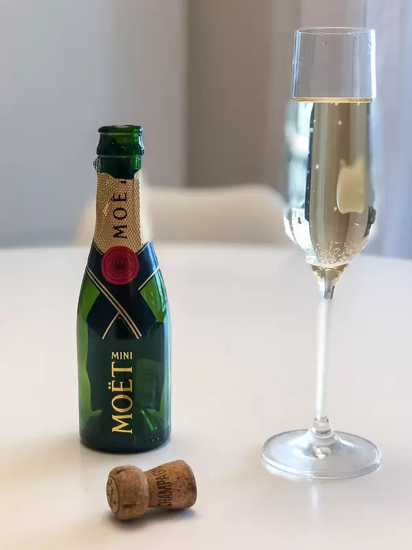 An empty bottle of Mini Moët, a full glass of Champagne and a cork on a white table