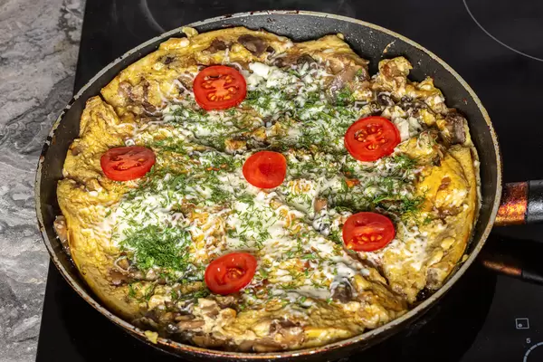 An omelet with mushrooms, cheese, tomatoes and herbs is fried in a frying pan