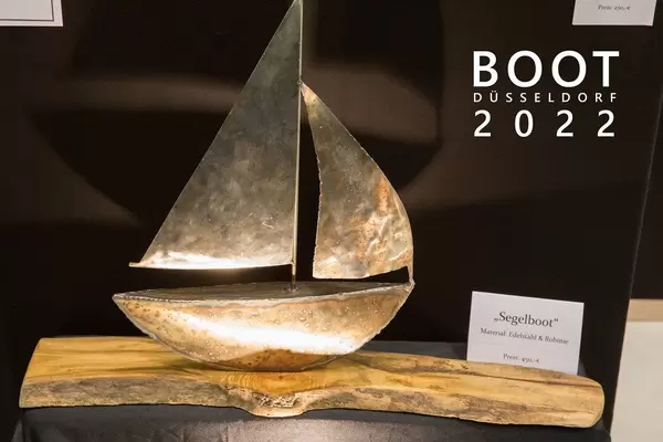 Angestrahltes Kunstwerk auf einem Holzstamm zeigt ein Segelboot aus Edelstahl und Robinie, neben dem Bildtitel "Boot Düsseldorf 2022"