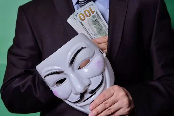 Anonymous government official with mask putting a stack of us dollars into his suit pocket. Concept of bribe, corruption in government
