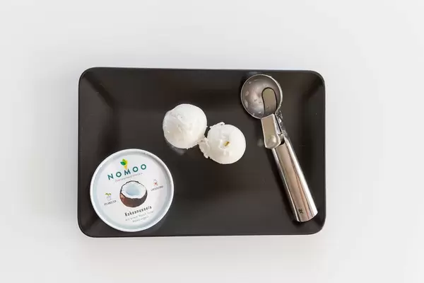 Ansicht von oben auf das Veggieeis Nomoo Kokosnuss mit Kokosraspeln, zwei Eiskugeln und einem Eislöffel auf einem schwarzen Teller