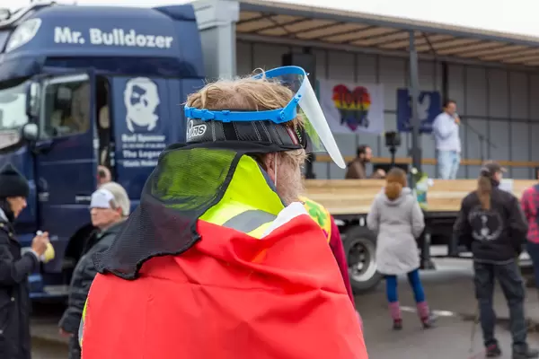 Anti-Corona-Demo in Köln: rauchender Mann trägt Gesichtsvisier, Warnschutzjacke und roten Mantel