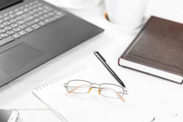 Arbeitstisch mit Utensilien wie Laptop, Schreiber, Notizheft, Smartphone, Kalenderbuch und Brille