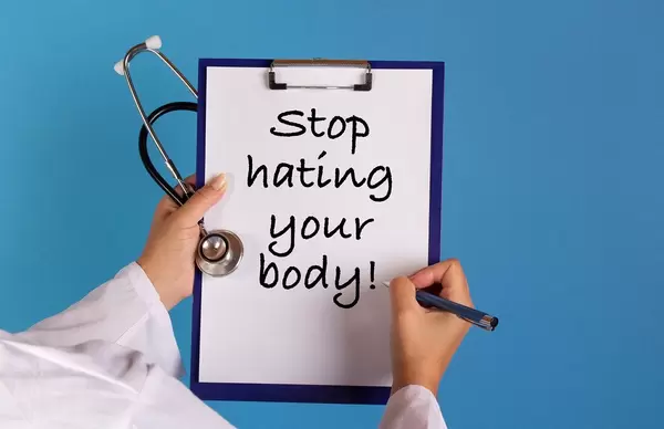 Arzt mit einem Stethoskop in der Hand hält ein Schild mit der Schrift 'Stop hating your body!'