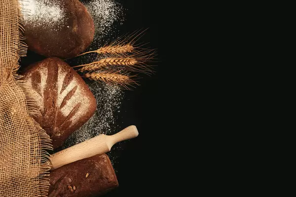 Assortment of black bread on a dark background with rolling pin, burlap and spikelets, top view
