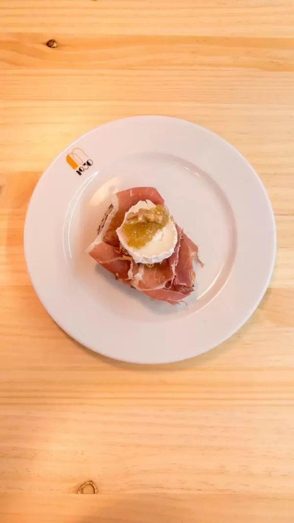 Aufsicht - Brot-Häppchen mit Serano Schinken, Weichkäse, Gelee und einer Walnuss auf einem weißen Teller