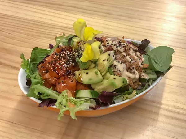Aufsicht - Ein Salat aus Baby-Spinat, Gurken, Rucola, Avocado, Tomaten, Dressing, Blüten und Sesam in einem ovalen Teller