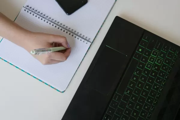 Aufsicht einer Frauenhand, schreibend auf Notizheft, mit Laptop und Smartphone