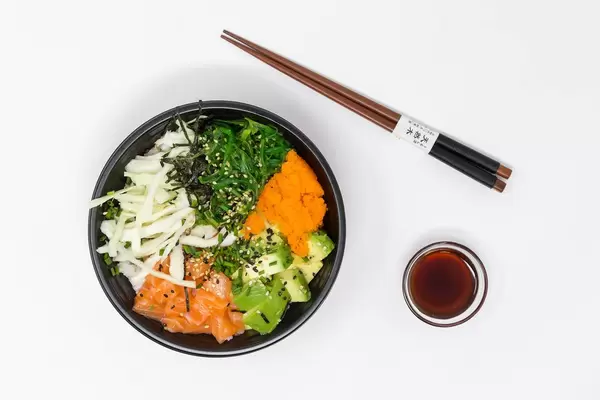 Aufsicht - Poke Bowl Lachs Teriyaki - mit Sushireis, Lachs, Avocado, Krautsalat, Wakame Salat, Wasago, Nori, Teriyaki-Sauce, Soja-Sauce, Sesam und Schnittlauch in einer Schüssel mit Essstäbchen und einer Schüssel Soya-Sauce