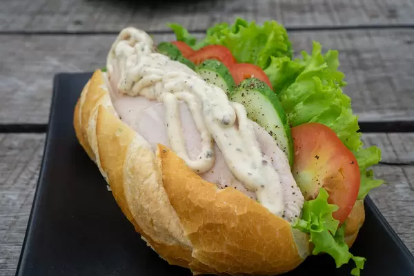 Baguette Sandwich mit gegrillter Hähnchenbrust, Gurke, Tomaten, Salat und Remoulade auf einem schwarzen Teller Nahaufnahme