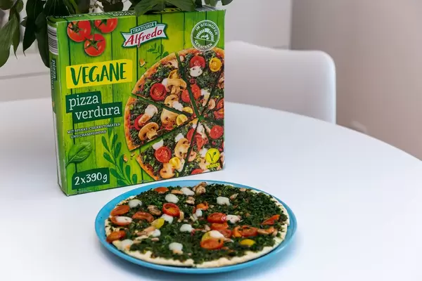 Baked pizza Verdura by Trattoria Alfredo with spinach topping, mushrooms and cherry tomatoes in front of the pizza box, on a white table