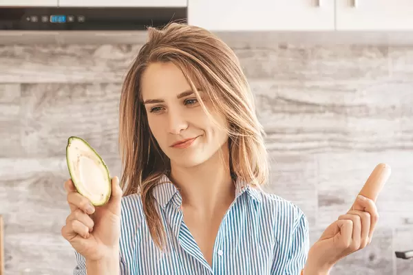 Beautiful girl in the kitchen with a sausage and avocado in her hands. The concept of choosing between healthy and unhealthy food