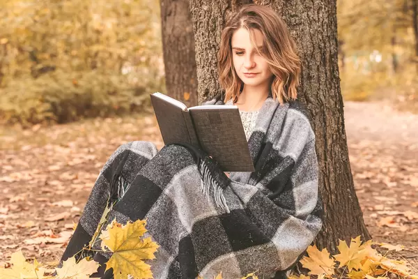 Beautiful young girl sitting on a fallen autumn leaves in a park, reading a book