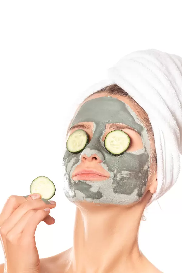 Beautiful young woman with clay mask on her face holding slices of fresh cucumber, skin care and treatment and cosmetology concept