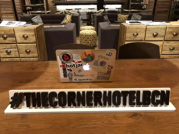 Beklebtes Macbook auf einem Holztisch in der Lounge vom The Corner Hotel, hinter dem Hashtagbanner #TheCornerHotelBCN