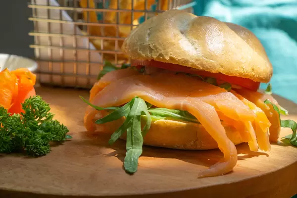 Belegtes Brötchen mit Räucherlachs, Rucola, Tomaten und Honig Senf Sauce auf einem Holzbrett mit Petersilie und einem kleinen Korb mit Pommes Frites im Hintergrund Nahaufnahme
