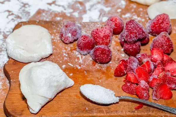 Bereitgestellte Zutaten für Teigtaschen mit Teig, gefrorenen Erdbeeren und Zucker auf Holzbrett