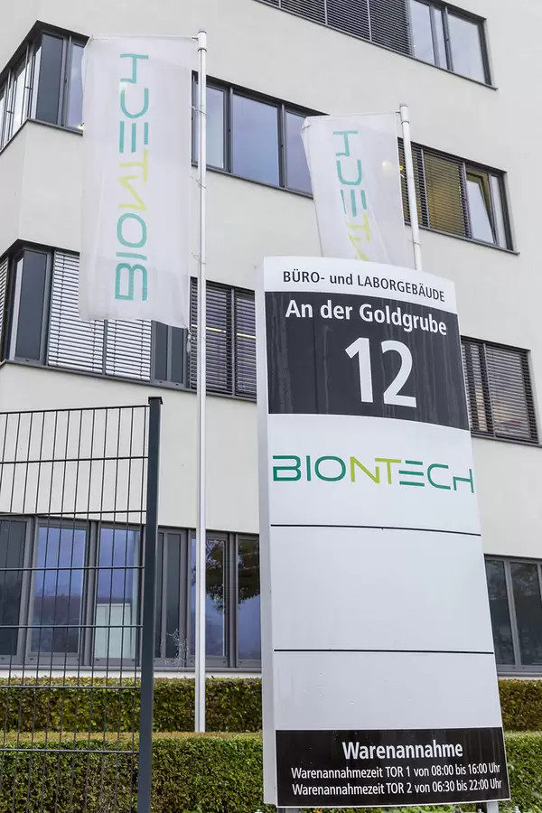 BioNTech Information Signboard with Address An der Goldgrube 12 showing Opening Hours of Acceptance of Goods with BioNTech Flags and Building in the Background