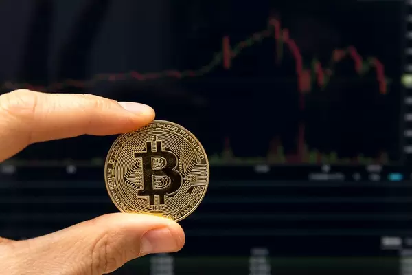 Bitcoin between to fingers in detail infront of falling stock prices on a display