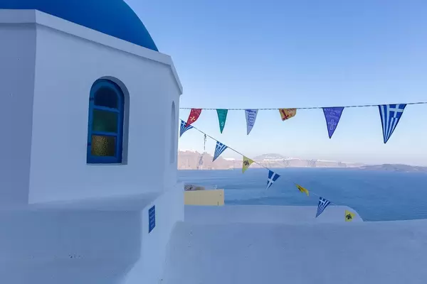 Blau und weiss auf Santorin: weißes Gebäude mit blauer Kuppel, Fahnen und dem Meer