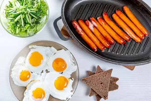 Blick von oben auf Frühstückstisch mit gebratenen Würstchen, Spiegeleiern, Toast und Salat