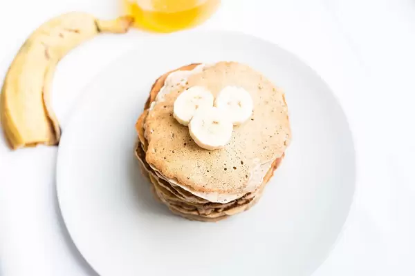 Blick von oben auf gestapelte, vegane Pfannkuchen mit Bananenstücken auf weißem Teller