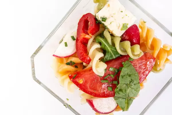 Blick von oben auf Glasschale mit Salat aus Nudeln, eingelegtem Käse, Tomate, Radieschen und Rucola