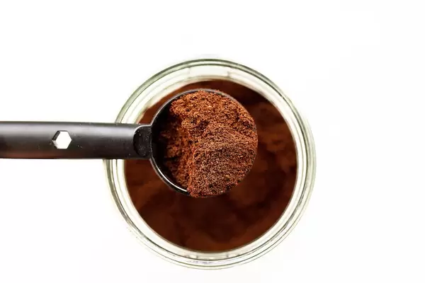Blick von oben auf Löffel mit fein gemahlenem Kaffeepulver über Glas mit Kaffee vor weißem Hintergrund