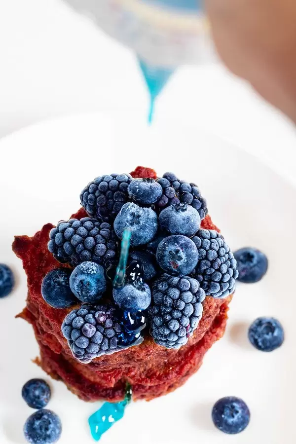 Blue topping pours on top of beetroot pancakes with blueberries and mulberries