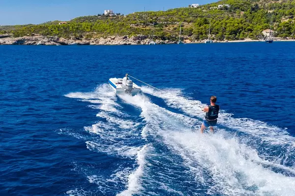 Blue-white water ski boat pulling a water sports man across the deep blue sea, in front of the rocky beach of Spetses, Greece