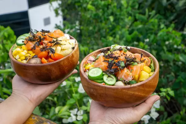Bokeh Food Photo of Person holding two Wooden Bowls with Healthy Hawaiian Poke Dish with Plants and Flowers in the Background