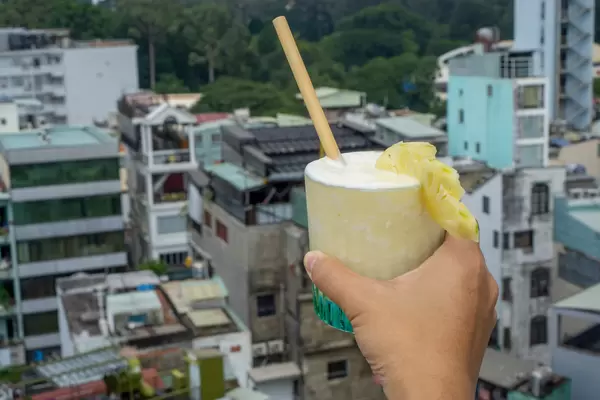 Bokeh Photo of Person holding a Virgin Pina Colada in a Cocktail Glas with Bamboo Straw and Slice of Pineapple with Buildings in the Background