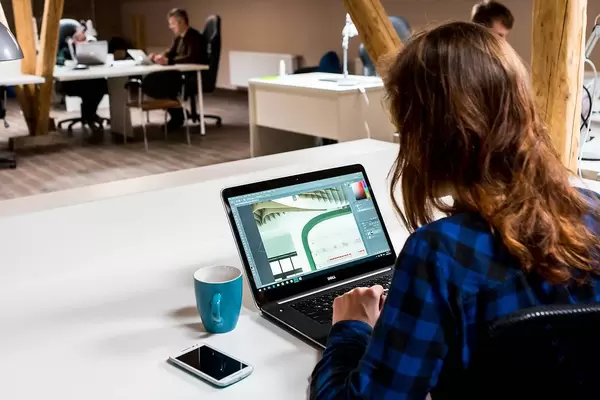 Bokeh Photo of Person working in an Office with Photoshop on Laptop with Phone and Coffee Cup next to it